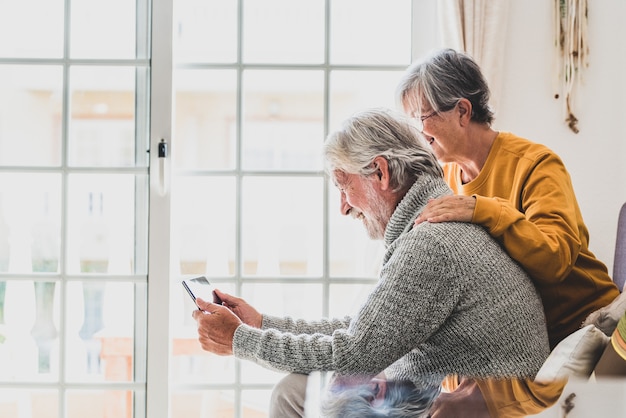 Couple of two cheerful and happy mature and old people seniors using tablet and having fun sitting on the sofa at home together. Beauty retired pensioners using internet and surfing the net