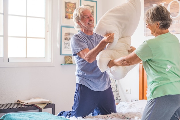 Coppia di due anziani felici che si divertono a giocare insieme sul letto di casa combattendo con i cuscini godendosi la guerra dei cuscini al chiuso al mattino