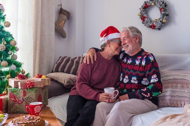 Couple of two happy seniors and mature people sitting in the sofa having fun together wrapping the gifts and presents - pensioners in love at home