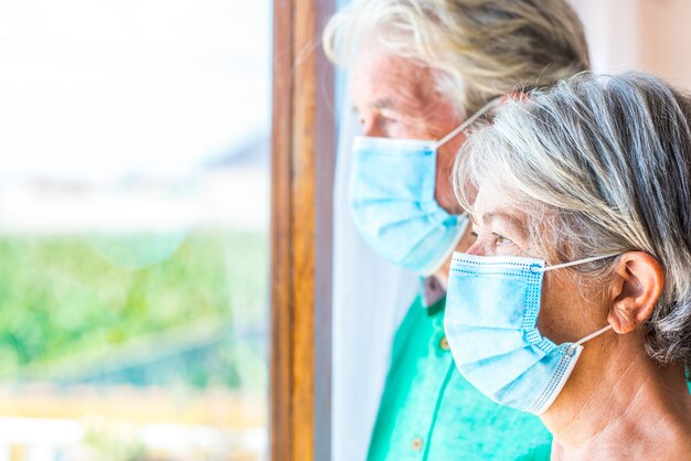 Photo couple of two senior and mature people looking outdoors from their home indoors wearing mask to prevent coronavirus - lockdown lifestyle