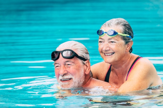 Couple of two seniors hugged in the water of swimming pool - active man and woman doing exercise together at the pool - hugged with love - wearing goggles