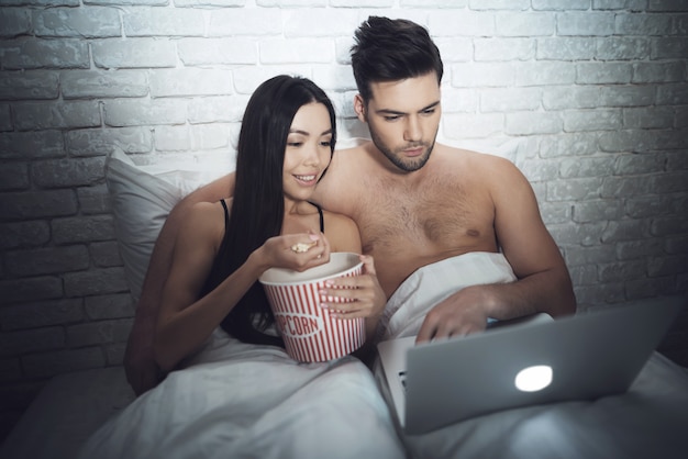 Пара с ноутбуком лежит на кровати и смотрит фильм