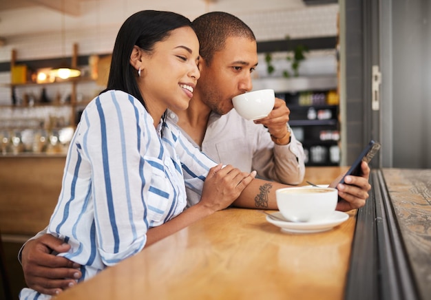 Couple using social media on a phone and drinking tea in a coffee shop together Happy man and woman with 5g mobile smartphone texting and searching on an online app and enjoy a date at a cafe