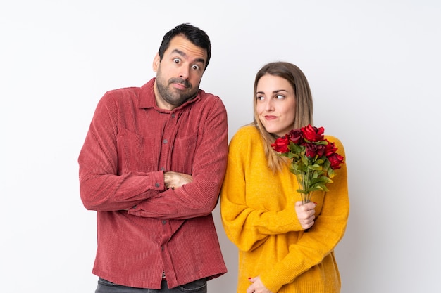 Couple in Valentine Day holding flowers over isolated wall making doubts gesture while lifting the shoulders