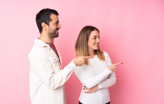 Couple in Valentine Day over isolated pink wall pointing finger to the side in lateral position