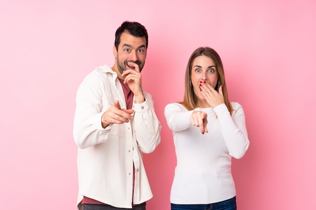 Couple in Valentine Day over isolated pink wall pointing with finger at someone and laughing