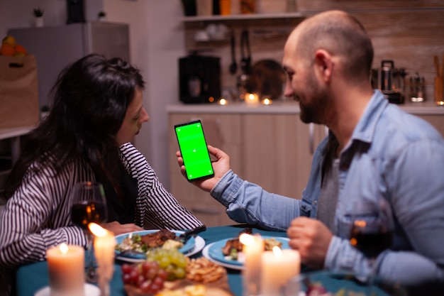 Couple watching phone with green screen. Happy man and woman looking at green screen template chroma key isolated smartphone display using technology internet sitting at the table in kitchen.