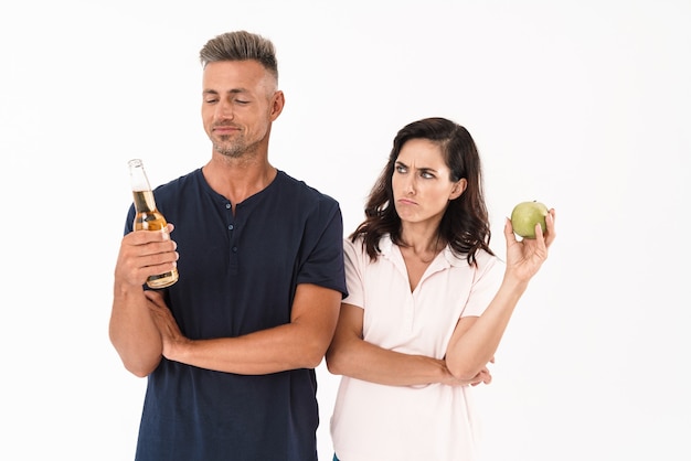 Coppia che indossa un abito casual in piedi isolato su un muro bianco, uomo felice con in mano una bottiglia di birra, donna arrabbiata con in mano una mela verde