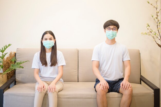 保護のためとコロナウイルスを避けるために、マスクをしているカップルが隔離中に自宅のソファーに座っています。