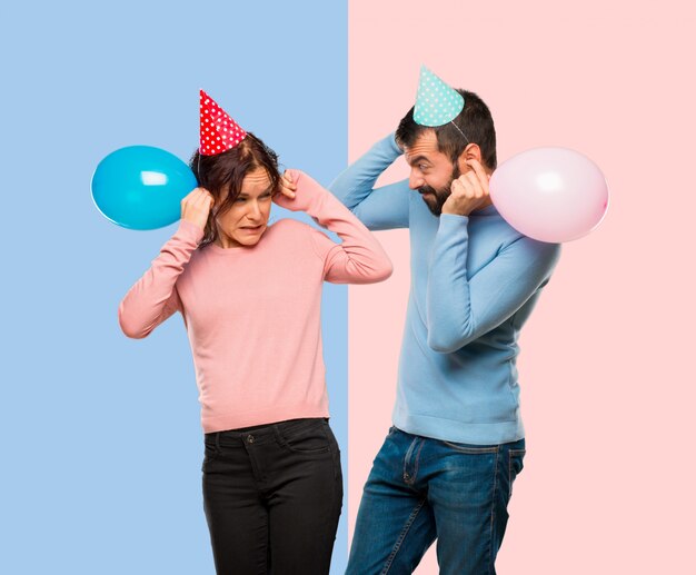 Couple with balloons and birthday hats covering ears with hands 