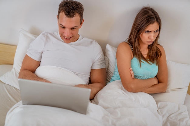 Coppia con laptop a letto. uomo soddisfatto che naviga in rete al computer portatile, donna sconvolta e arrabbiata guardando dall'altra parte.