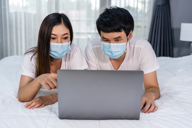 Le coppie con le maschere mediche che per mezzo del computer portatile sul letto durante la quarantena pandemia coronavirus restano a casa.