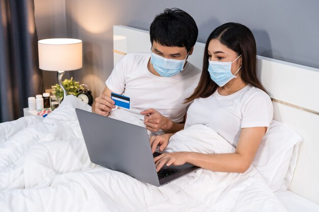 Le coppie con le maschere mediche che per mezzo del computer portatile per comperare online con la carta di credito sul letto durante la quarantena il coronavirus pandemico restano a casa.