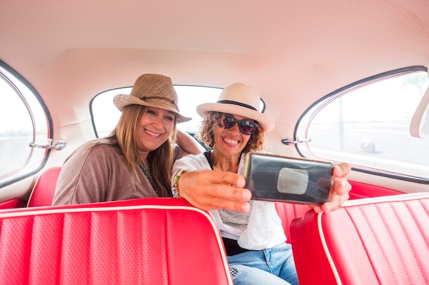 couple of women caucasian friends enjoying a red vintage car taking selfie with a modern smartphone