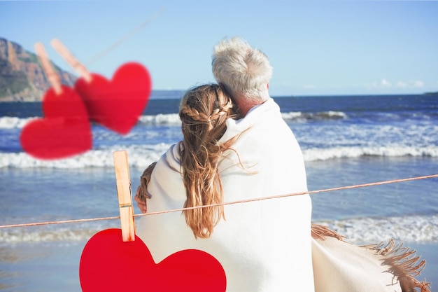 Пара, завернутая в одеяло на пляже, глядя на море против сердец, висящих на веревке