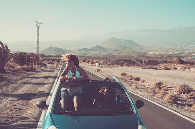 Couple of young adult women on a convertible car driving a long road with desert