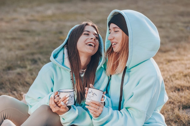 A couple of young attractive girls dressed in identical blue hoodies are sitting on the grass, drinking tea and laughing. Socializing with friends in nature.