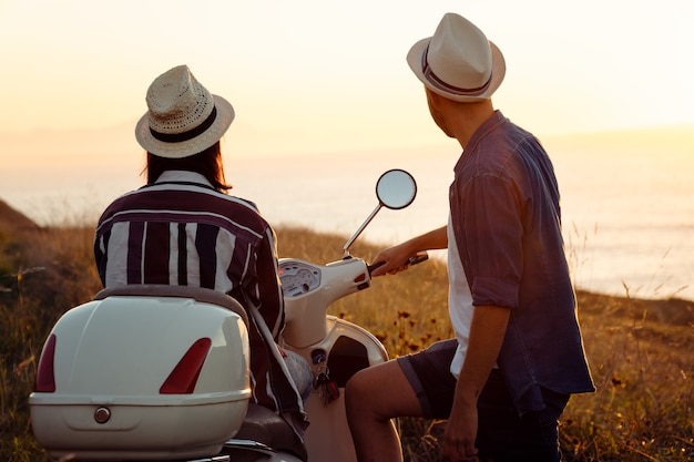 新鮮な服と帽子をかぶった若い友人のカップル、夕日の光の中で海岸でバイクに乗って楽しんで笑っている