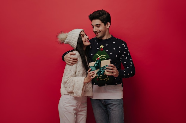 Couple of young people in cozy knitted sweaters and jeans with Christmas gift box smiling looking to each other and posing against red background