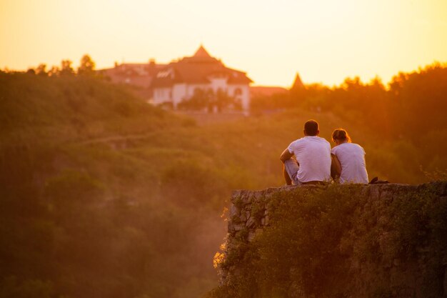 A couple of young people sit on the edge of a cliff as the sunset