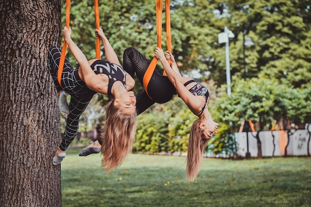 Couple of young sportive womans is doing exercises upside down under big tree in the green park.