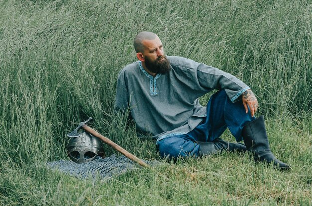 Photo courageous man with beard resting in grass in field pensive gaze next to him is ax warrior's helmet