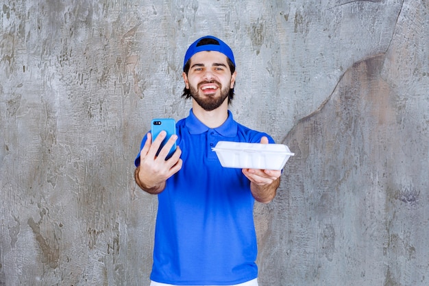 Courier in blue uniform holding a plastic takeaway box and taking new orders via phone. 