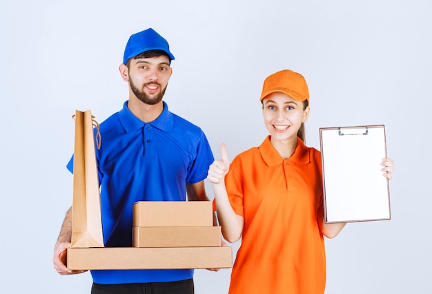 Courier boy and girl in blue and yellow uniforms holding cardboard takeaway boxes and shopping packages, presenting signature list and feeling satisfied.