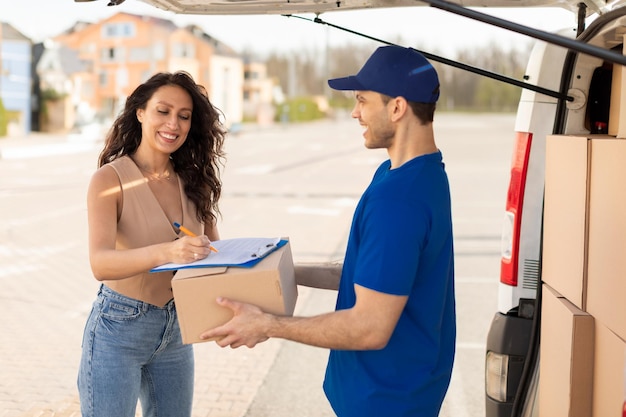 Foto courier delivery concept happy young woman receiving parcel and signing order receipt outdoors