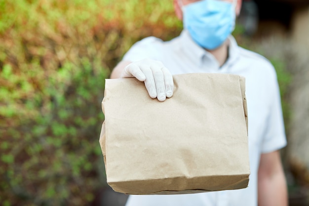 Corriere, fattorino in guanti medici in lattice consegna in sicurezza acquisti online in sacchetti di carta marrone alla porta durante l'epidemia di coronavirus