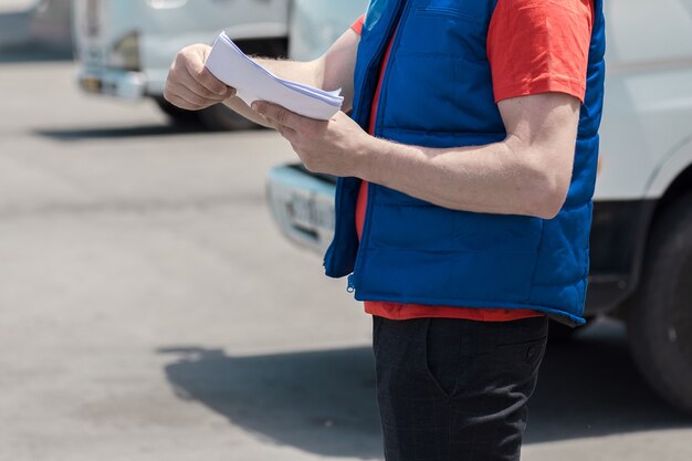 制服を着た宅配便の運転手が、彼の後ろにある書類と配達用の白いトラックにメモをとっています。