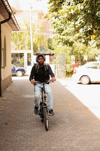 Courier in headphones riding bike on street at sunny day, looking at camera, front view. African american deliveryman with customer order in thermal bag portrait, man delivering eatery meal