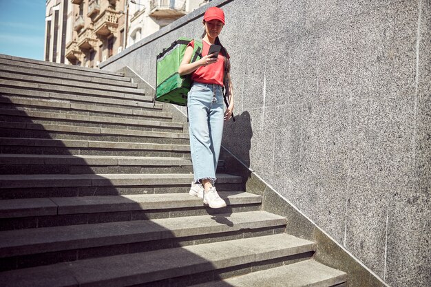 Courier lady with thermal insulated bag for food looking for location using mobile phone while delivering order in urban area