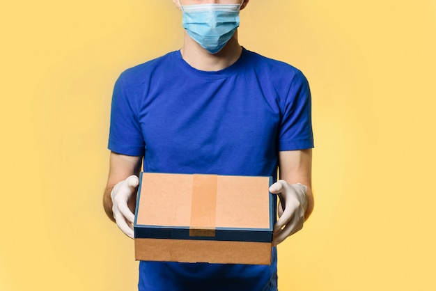 A courier in a medical mask and protective gloves holds a parcel, standing isolated on a yellow wall with place for text.