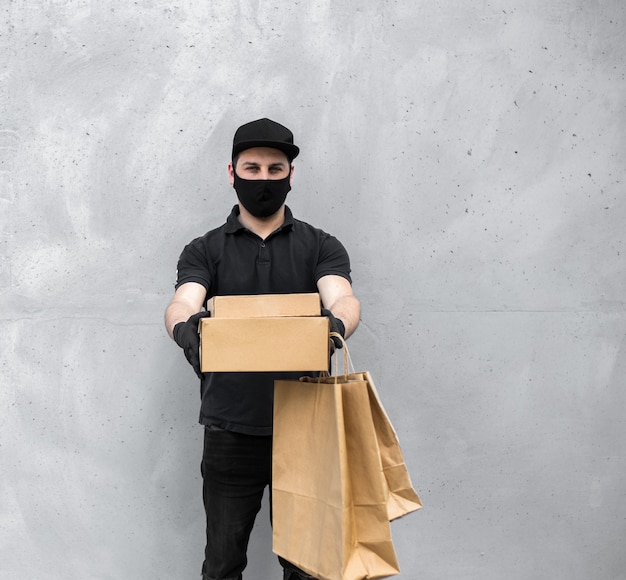 Courier in protective mask and medical gloves delivers takeaway food near grey wall