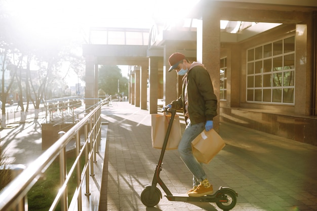 Courier in protective mask and medical gloves on a scooter delivers craft paper packet with food. Delivery service under quarantine, disease outbreak, coronavirus covid-19 pandemic conditions.