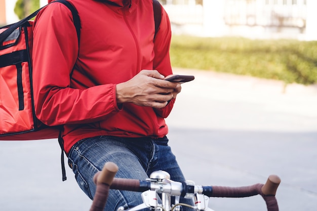 Corriere in uniforme rossa con una scatola di consegna sul retro in sella a una bicicletta e guardando il cellulare per controllare l'indirizzo per consegnare il cibo al cliente. corriere in bicicletta che consegna cibo in città.