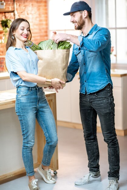 Courier service worker delivering fresh food, giving shopping bag to a happy woman client on the kitchen at home