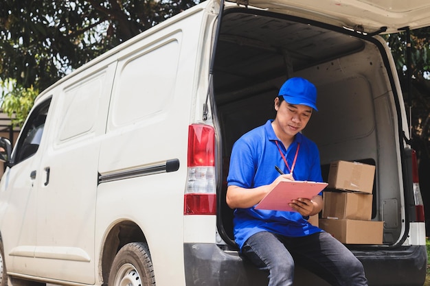 Photo courier sitting beside the box package and checking list shipment on the trunk of the van
