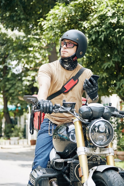 Courier with smartphone in hands sitting on motorcycle and waiting for customer to come down to take...