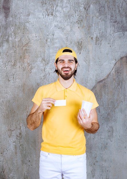 Courier in yellow uniform holding a disposable cup and introducing business card. 