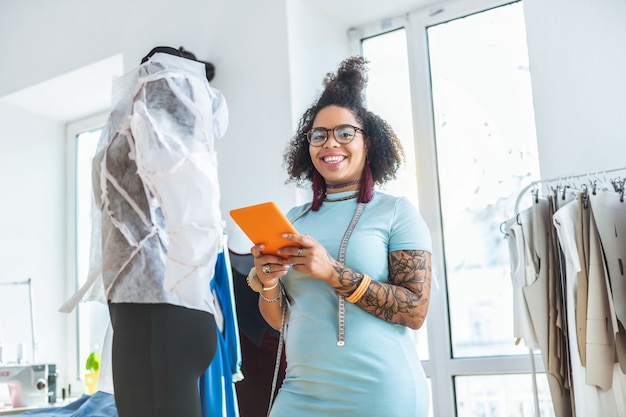 Coperto di tatuaggi. donna allegra sorridente che indossa abito blu e trascorrere la giornata in studio attrezzato per la produzione di vestiti