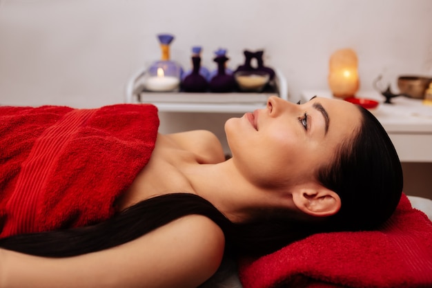 Covered with towel. Appealing dark-haired female with ponytail calmly resting in massage cabinet and waiting for procedure