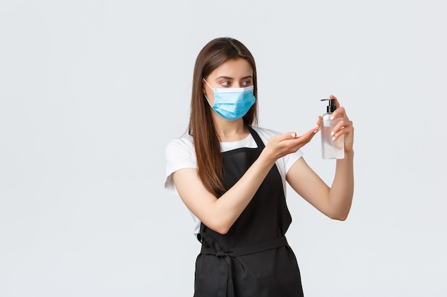 Covid-19, сотрудники кафе, кафе и концепция коронавируса. Бариста в медицинской маске и черном фартуке использует дезинфицирующее средство для рук на работе, чтобы предотвратить вирус во время пандемии, соблюдая личную гигиену.