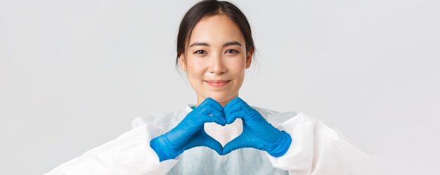 Covid-19, coronavirus disease, healthcare workers concept. Close-up of exhausted happy asian female doctor take-off personal protective equipment, have skin marks from respirator, show heart gesture.