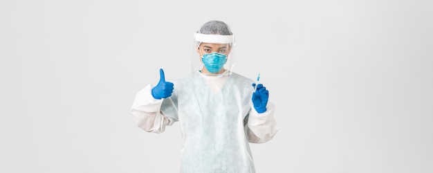 Covid-19, coronavirus disease, healthcare workers concept. Confident serious female asian doctor in personal protective equipment, show thumb-up and hold syringe with vaccine, white background.