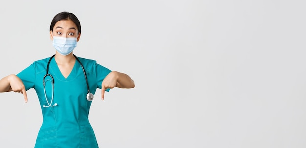 Covid-19, coronavirus disease, healthcare workers concept. Excited and surprised asian female physician, nurse in scrubs and medical mask, pointing fingers down, showing advertisement.