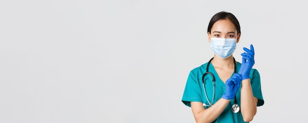 Covid-19, coronavirus disease, healthcare workers concept. Professional smiling asian female physician, doctor in medical mask and scrubs, put on rubber gloves ro examination, white background.