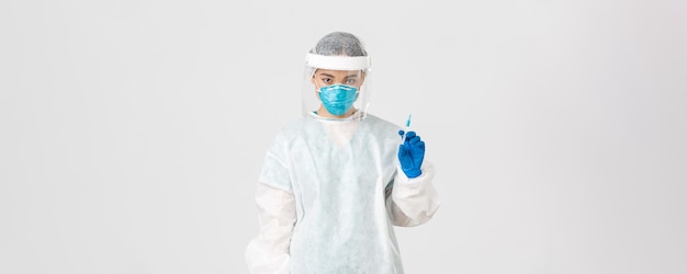 Covid-19, coronavirus disease, healthcare workers concept. Serious-looking confident asian female doctor in medical respirator and personal protective equipment, holding syringe with vaccine