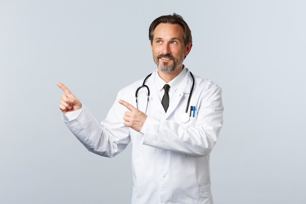 Covid-19, coronavirus outbreak, healthcare workers and pandemic concept. Intrigued male doctor reading sign, pointing fingers upper left corner at banner promo, wearing white coat